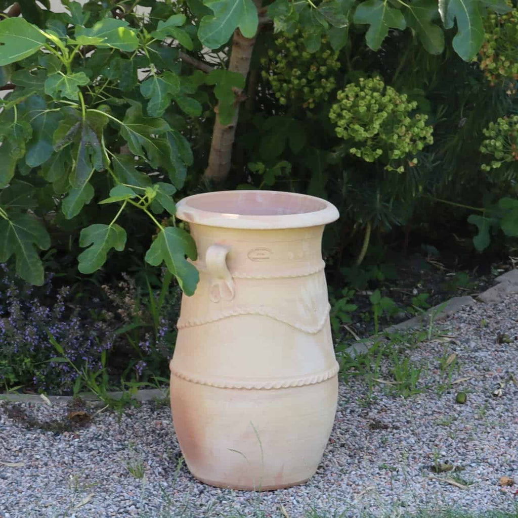 Eine hohe, beige Keramikvase mit dekorativen Mustern, die der griechischen schmalen Terrakotta-Amphore „Cistus“ (H=60 cm) ähnelt, steht auf einem Kiesweg neben einem üppigen grünen Busch in einem Garten. Die Vase hat einen Griff und ist von kleinen Pflanzen und Grün umgeben.