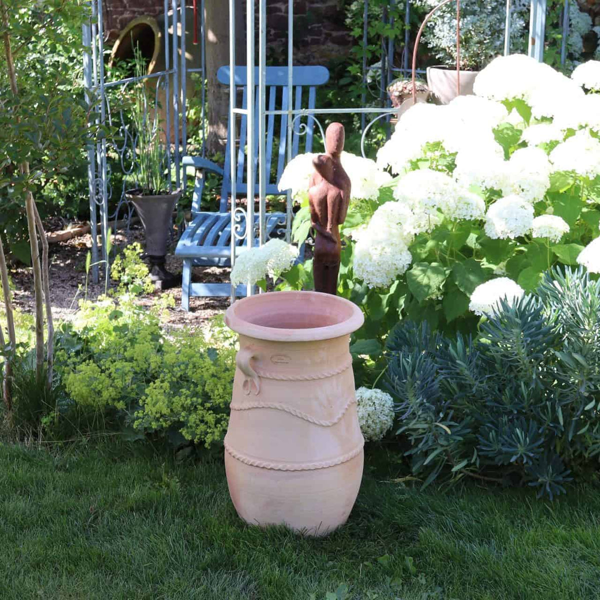 Eine Gartenszene mit einer großen, griechischen, schmalen Terrakotta-Amphore „Cistus“ H=60 cm auf dem Rasen. Der üppige Garten ist voller grüner Pflanzen und blühender weißer Cistus-Blüten. Neben der Amphore steht eine rustikale Holzstatue und im Hintergrund stehen blaue Gartenstühle aus Schmiedeeisen.