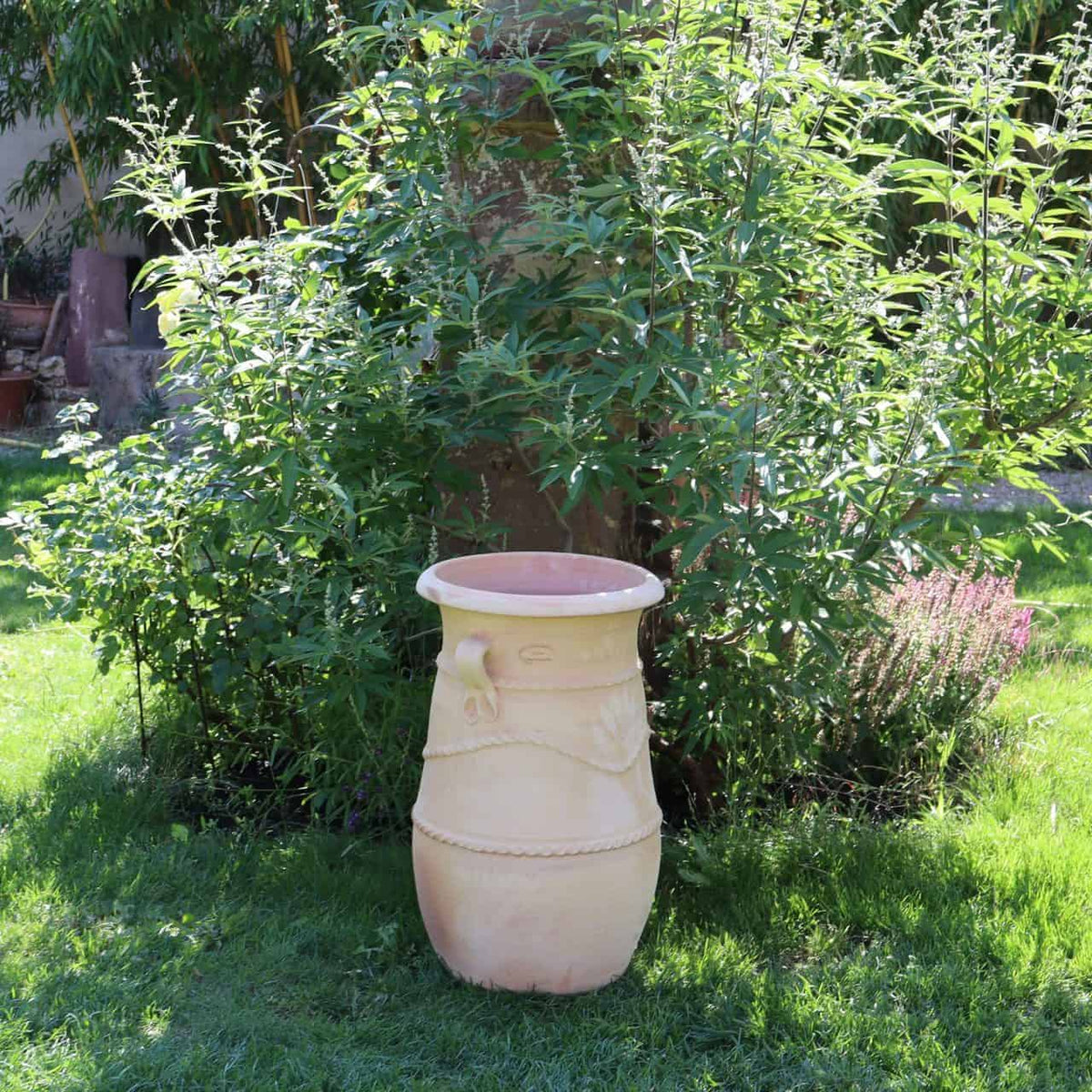 Die griechische schmale Terrakotta-Amphore „Cistus“ ist eine 60 cm hohe, große beige Keramikvase mit dekorativen Mustern, die an eine antike griechische Amphore erinnern. Auf dem Gras platziert und von üppigen grünen Büschen und Pflanzen in einem Garten umgeben, beleuchtet das Sonnenlicht sowohl die Vase als auch ihre grüne Umgebung und schafft eine lebendige und heitere Szene.