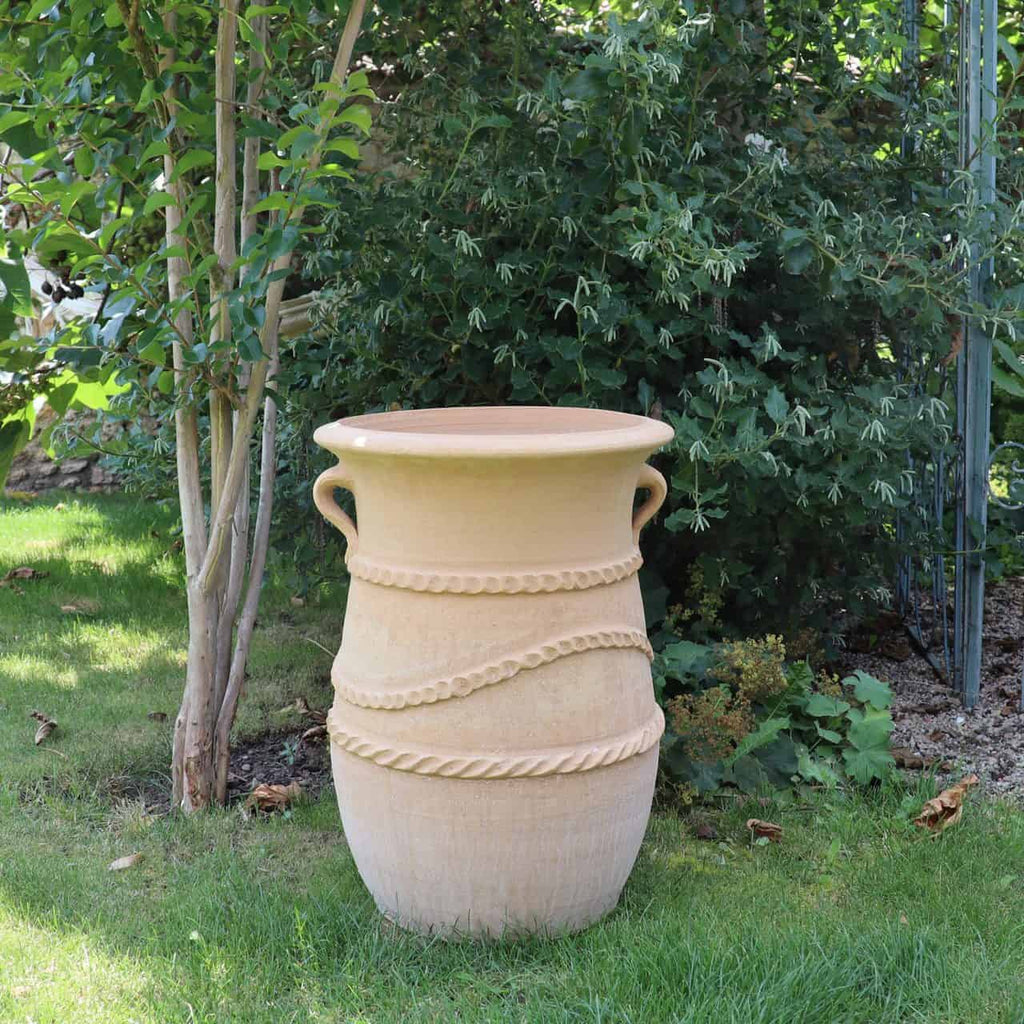 Eine große Terrakotta-Urne, genannt „griechische schmale Terrakotta-Amphore ‚Cistus‘ H=60 cm“, die einer griechischen Amphore mit dekorativen seilartigen Mustern um ihren Körper ähnelt, steht auf grünem Gras neben einem hohen Baum und belaubten Büschen. Die Urne hat zwei Griffe auf gegenüberliegenden Seiten. Sonnenlicht beleuchtet die Szene teilweise und hebt die Textur der Urne hervor.
