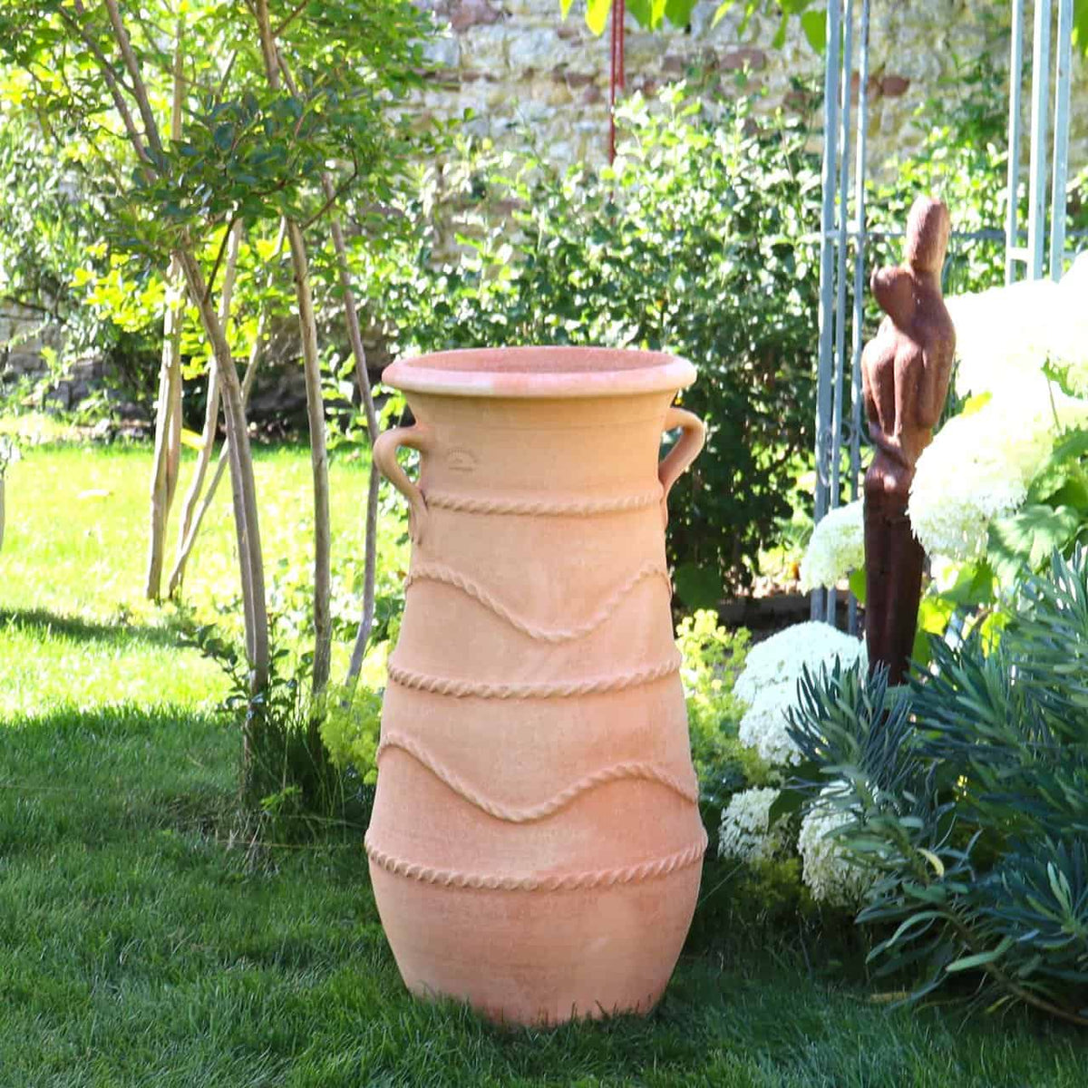 Eine große griechische Amphorenvase „Cistus“ H=80cm mit seilartigen Mustern steht auf einer Rasenfläche. Sie ist von üppigem grünem Laub, weißen Cistusblüten und Skulpturen umgeben, im Hintergrund ist teilweise eine Steinmauer zu sehen.
