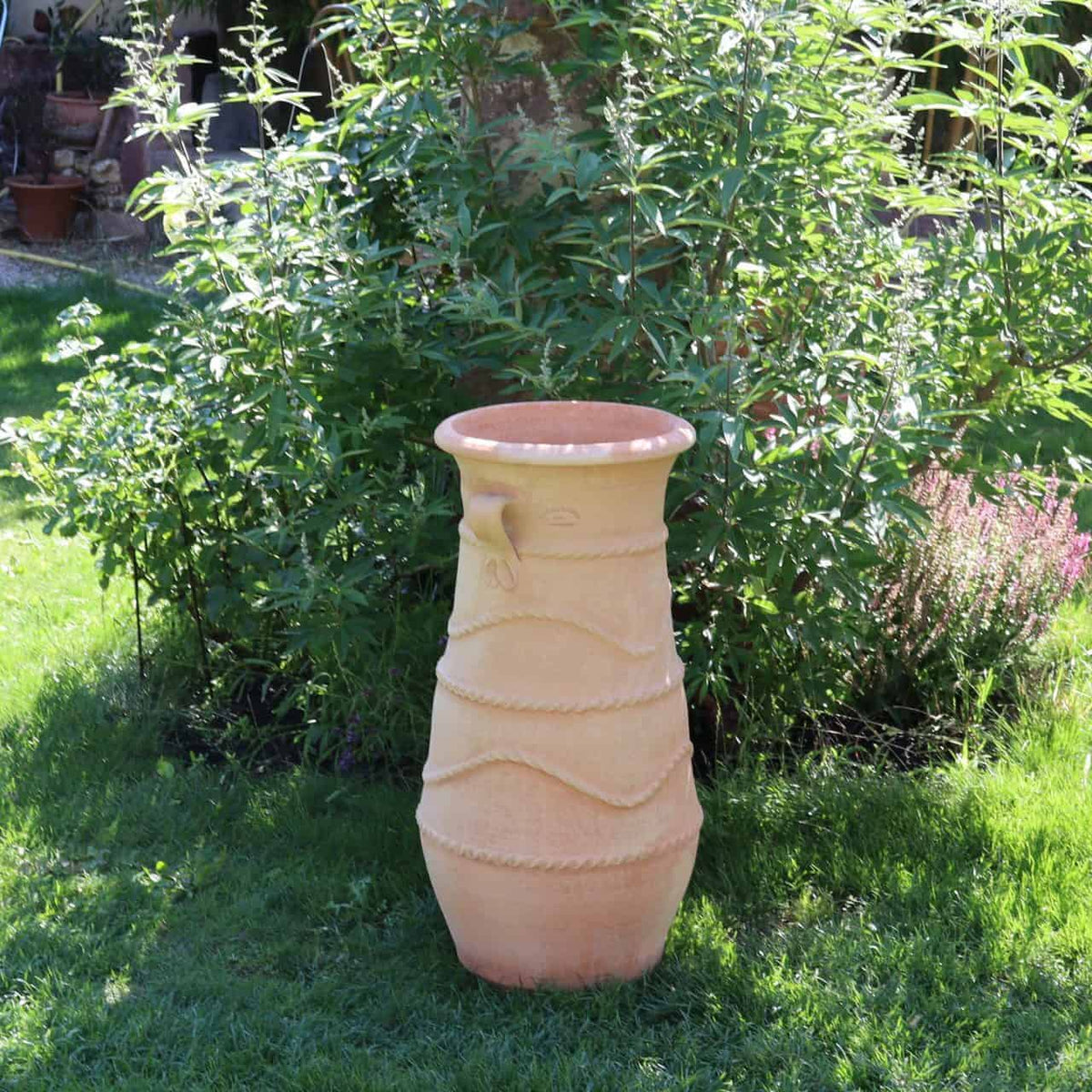 Die griechische Amphorenvase „Cistus“ H=80cm, groß und hellbraun mit dekorativen horizontalen Rillen und einem kleinen Ausguss oben, steht auf einer Rasenfläche, umgeben von üppigem grünem Laub. Im Hintergrund sind verschiedene Pflanzen und Cistus-Sträucher zu sehen, die eine lebendige Gartenszene schaffen.