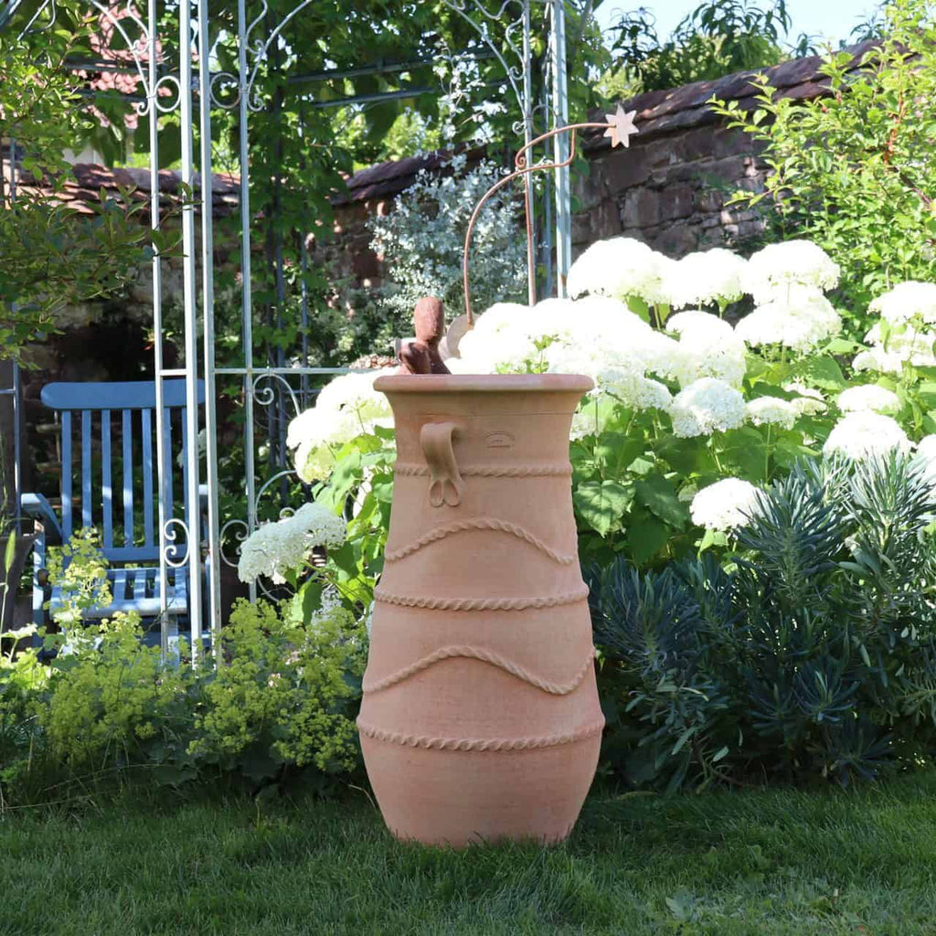In einem üppigen Garten, umgeben von Grünpflanzen und Zistrosen, steht eine hohe, dekorative Terrakotta-Urne, bekannt als griechische Amphorenvase „Cistus“ H=80cm, prominent. Im Hintergrund ist ein blauer Stuhl zu sehen und ein Metallgitter verleiht der Szene Struktur. Über der Urne befindet sich ein schmiedeeisernes Gartenornament mit Sternspitze.