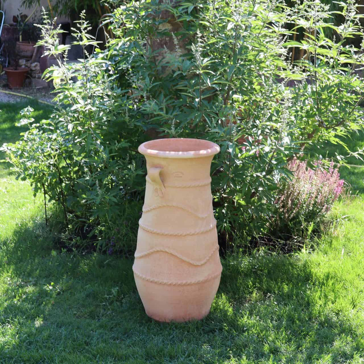 Eine große, hellbraune griechische Amphorenvase „Cistus“ (H=80cm) mit strukturierter Oberfläche steht auf einem üppigen grünen Rasen. Im Hintergrund sind dichte, belaubte Büsche und ein kleiner Cistus-Strauch mit rosa Blüten zu sehen, die eine ruhige Gartenszene schaffen. Sanftes Sonnenlicht erhellt die Vase und das umgebende Laub.