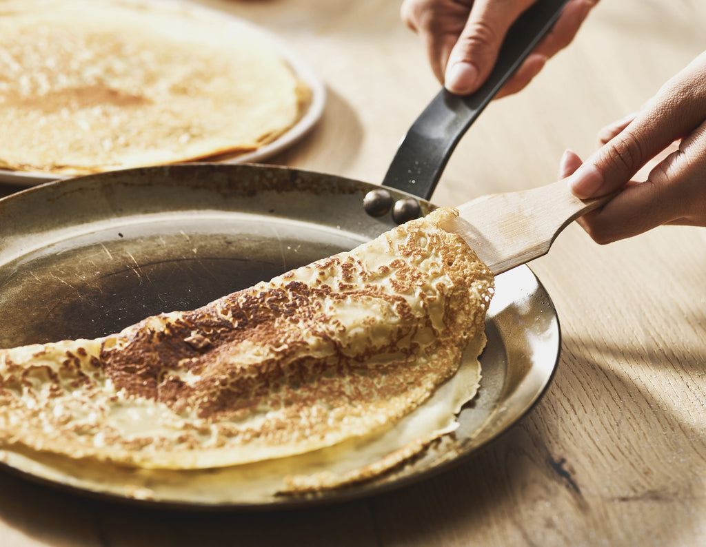 Nahaufnahme von Händen, die einen goldbraunen Crêpe mit einem Spatel in einer De Buyer Crêpe-Pfanne Carbone Plus auf einer hölzernen Oberfläche falten, wobei im Hintergrund ein anderer Crêpe zu sehen ist.