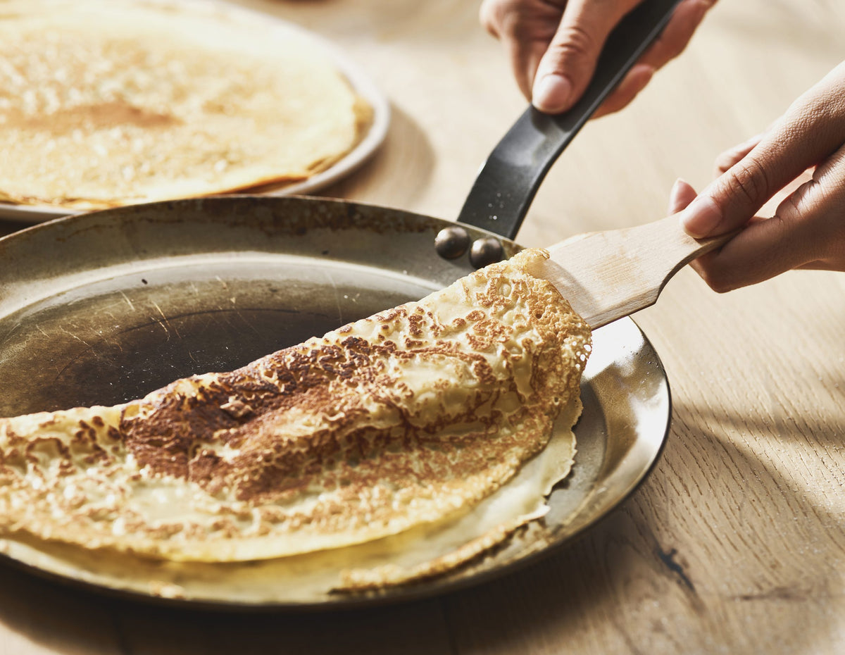 Nahaufnahme von Händen, die einen goldbraunen Crêpe mit einem Spatel in einer De Buyer Crêpe-Pfanne Carbone Plus auf einer hölzernen Oberfläche falten, wobei im Hintergrund ein anderer Crêpe zu sehen ist.