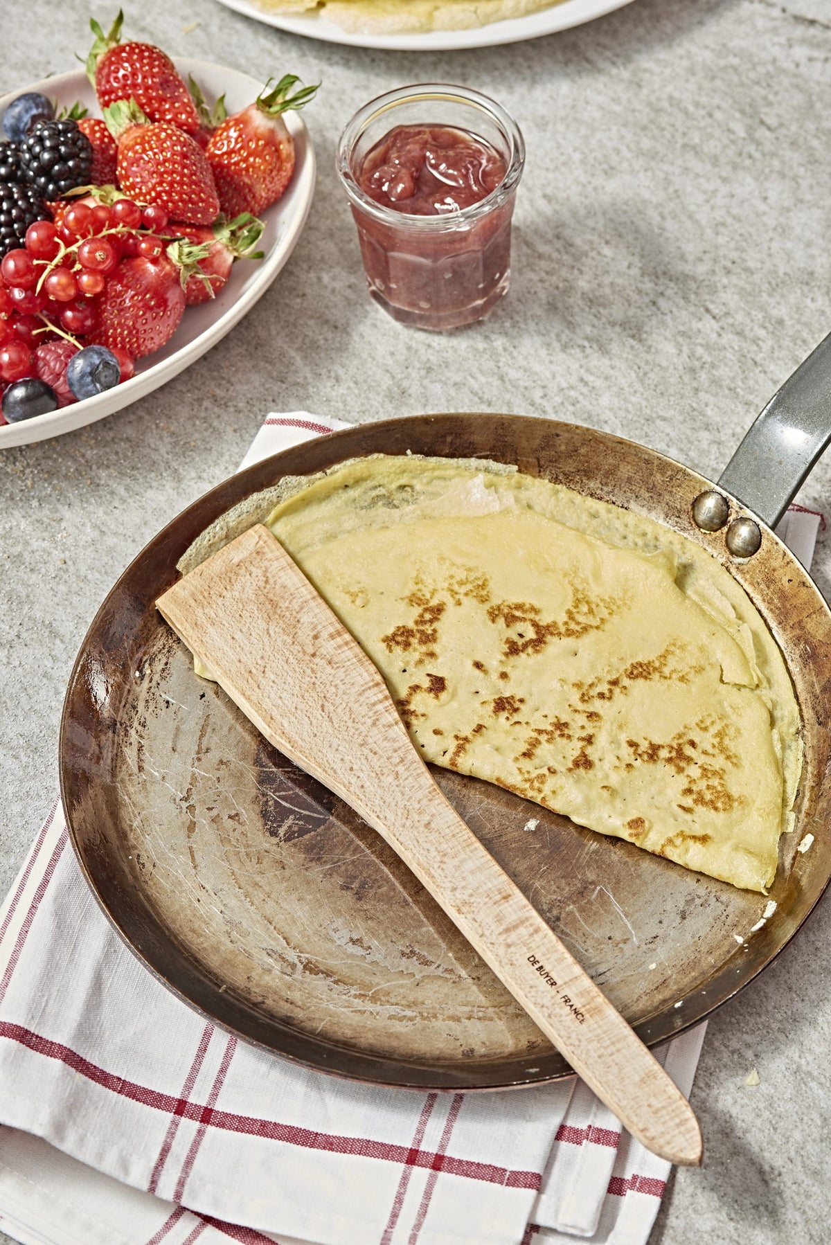 Eine De Buyer Crêpe-Pfanne Carbone Plus mit einem teilweise gegarten Crêpe und einem Holzspatel liegt auf einem gestreiften Tuch; daneben stehen ein Glas Marmelade und ein Teller mit verschiedenen frischen Beeren auf der grauen Fläche.