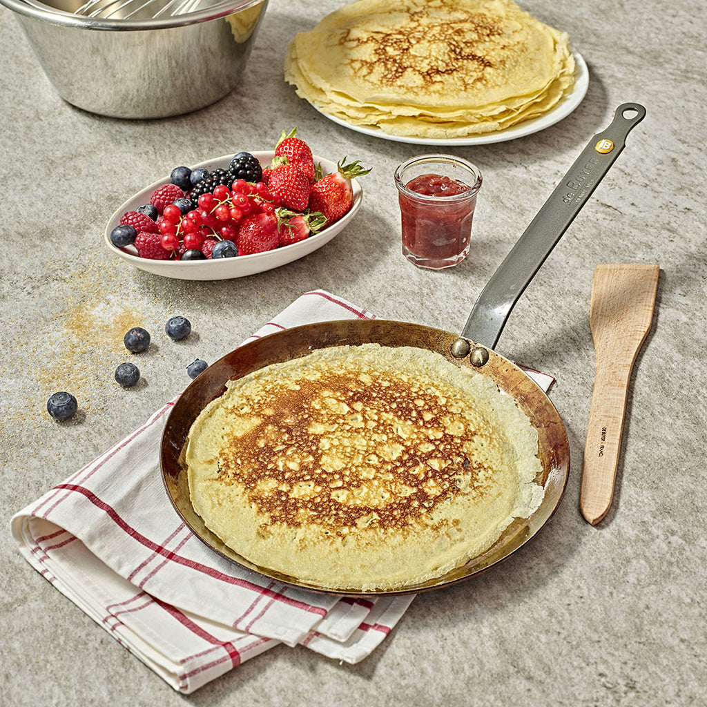 Ein gekochter Crêpe ruht in einer De Buyer Crêpe-Pfanne Mineral B, 26 cm Dm, auf einem gefalteten Handtuch, mit gestapelten Crêpes, gemischten Beeren, Marmelade in einem Glas, losen Blaubeeren und einem Holzspatel auf einer grauen Arbeitsplatte.