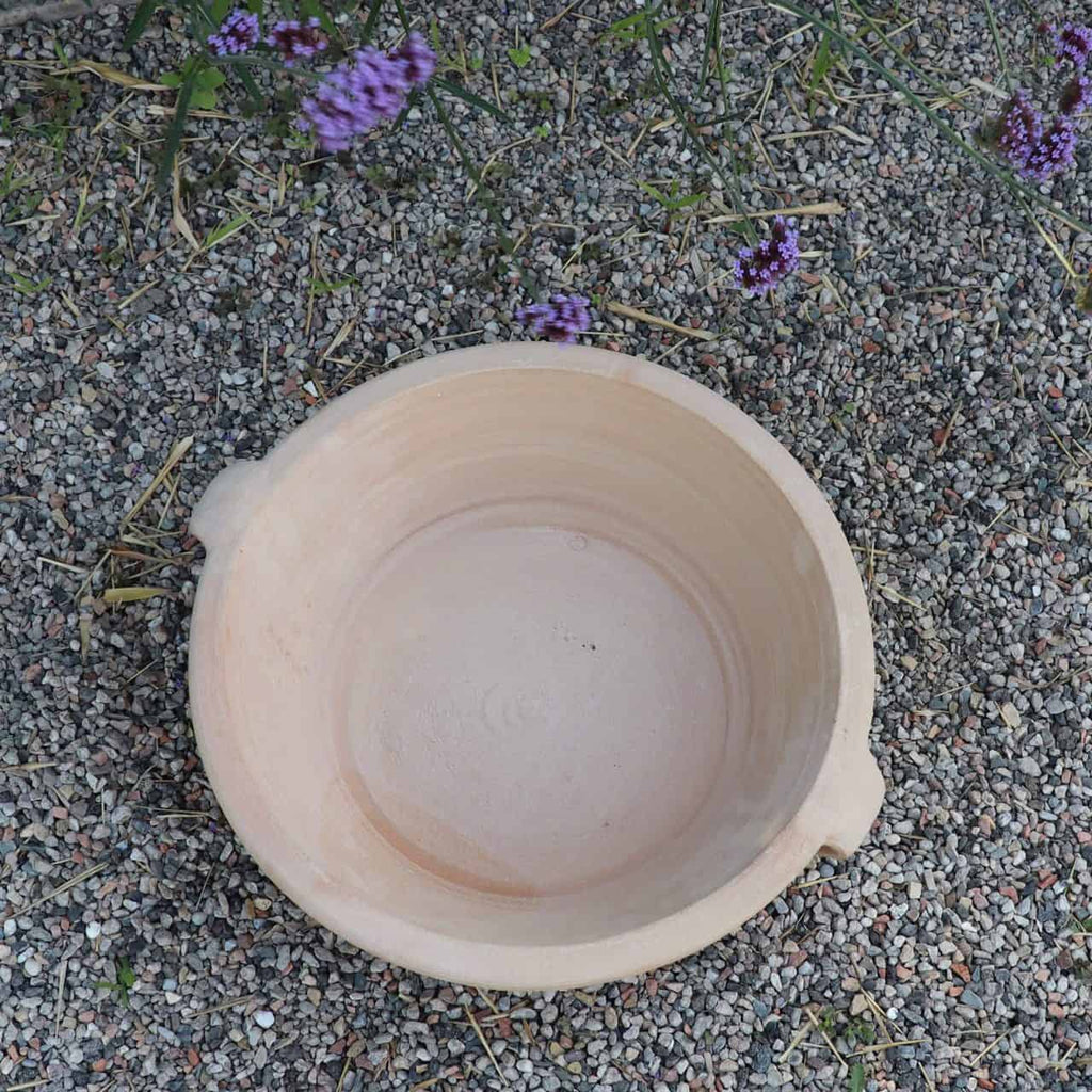 Eine große, leere Terrakotta-Pflanzschale „Krokus“ mit einem Durchmesser von 40 cm steht auf einer Kiesfläche, umgeben von spärlichem Laub. Am oberen und linken Bildrand ragen einige kleine violette Krokusblüten mit grünen Stielen hervor.
