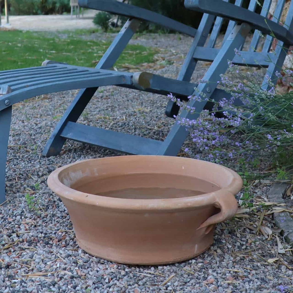 Eine große Terrakotta-Pflanzschale „Krokus“ mit 40 cm Durchmesser, gefüllt mit Wasser, steht auf Kies neben zwei blauen Holzliegestühlen. Im Hintergrund sind Lavendelblüten und Krokusblüten zu sehen, die den Charme der Gartenszene im Freien noch verstärken.