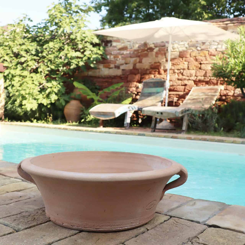 Ein ruhiger Außenpoolbereich bietet zwei Liegestühle unter einem weißen Sonnenschirm. Eine 40 cm große Terrakotta-Pflanzschale „Crocus“ steht im Vordergrund auf dem steinernen Pooldeck und verleiht ihm eine erdige Note. Üppiges Grün mit blühenden Krokusblüten und eine rustikale Ziegelwand verstärken das ruhige Ambiente.