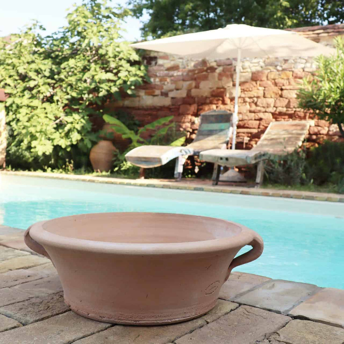 Ein ruhiger Außenpoolbereich bietet zwei Liegestühle unter einem weißen Sonnenschirm. Eine 40 cm große Terrakotta-Pflanzschale „Crocus“ steht im Vordergrund auf dem steinernen Pooldeck und verleiht ihm eine erdige Note. Üppiges Grün mit blühenden Krokusblüten und eine rustikale Ziegelwand verstärken das ruhige Ambiente.