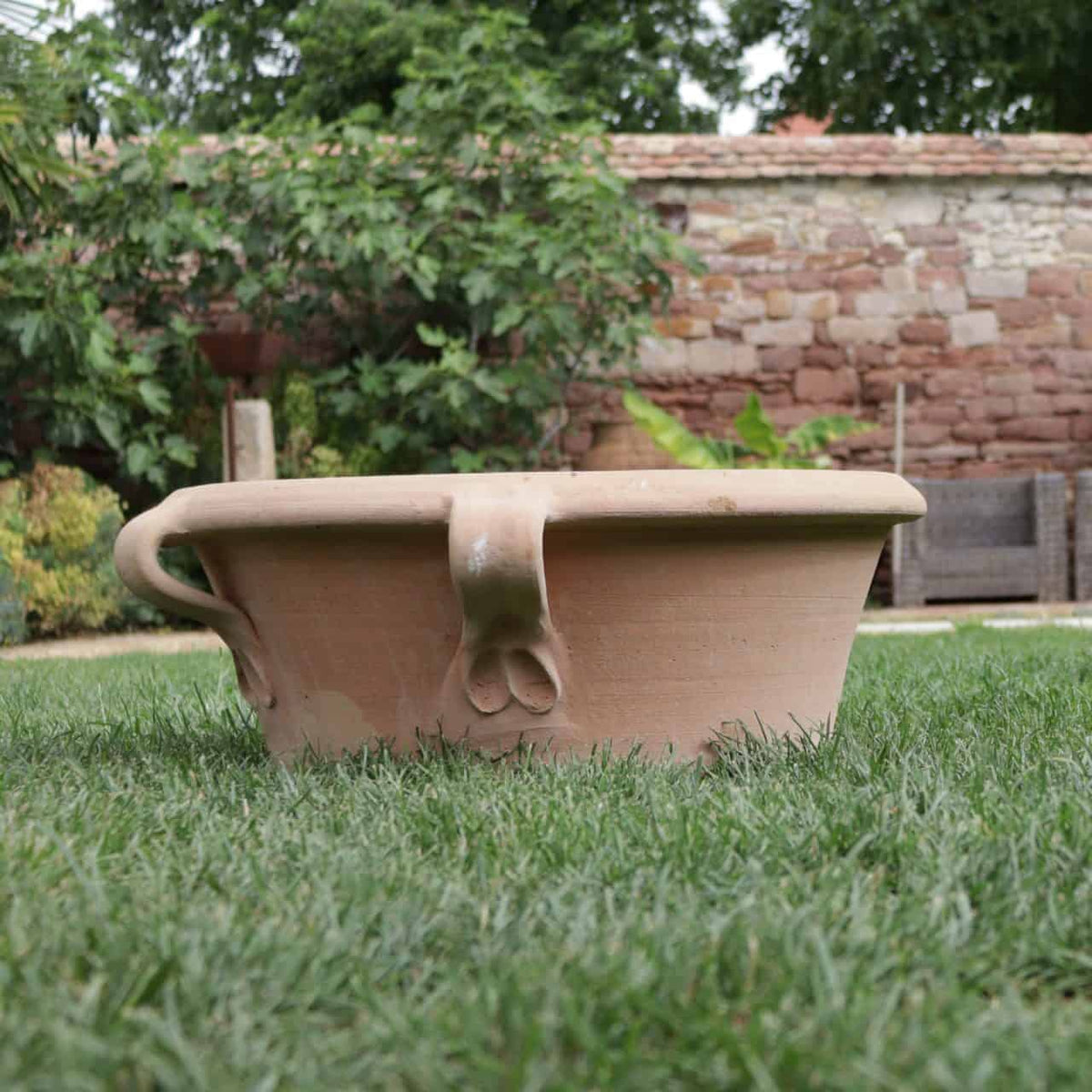 Eine niedrige, breite Pflanzschale „Crocus“ aus Terrakotta von Kreta Keramik mit zwei geschwungenen Griffen steht auf einem üppigen grünen Rasen. Im Hintergrund werten Sträucher, Bäume und eine rustikale Mauer aus Ziegeln und Steinen die Gartenkulisse auf. Dieses Stück verleiht dem ruhigen Außenbereich zeitlosen Charme.