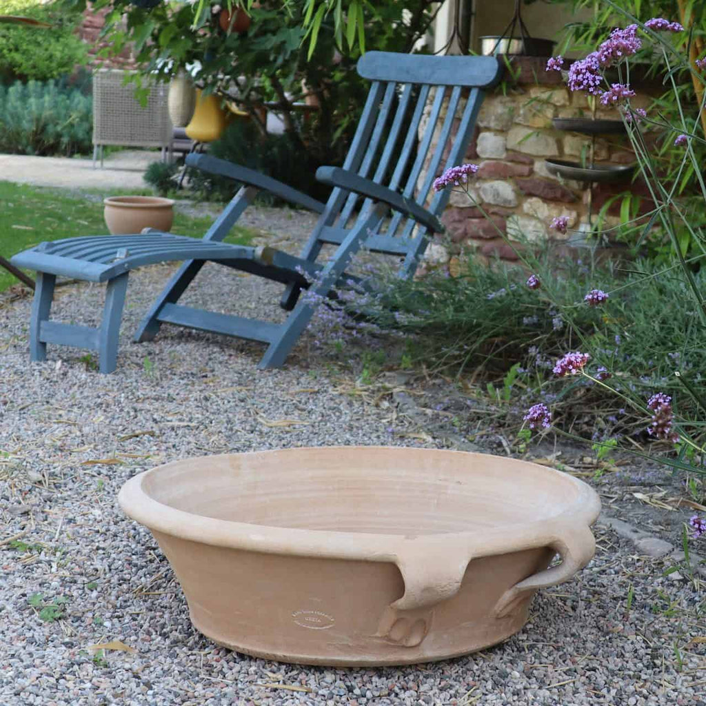 Eine ruhige Gartenszene zeigt einen blauen Liegestuhl aus Holz und eine passende Fußstütze auf Kies. Eine große, 40 cm große Terrakotta-Pflanzschale „Crocus“ mit integrierten Griffen steht im Vordergrund, umgeben von blühenden Krokussen und anderen violett blühenden Pflanzen, während im Hintergrund teilweise eine Steinmauer sichtbar ist.