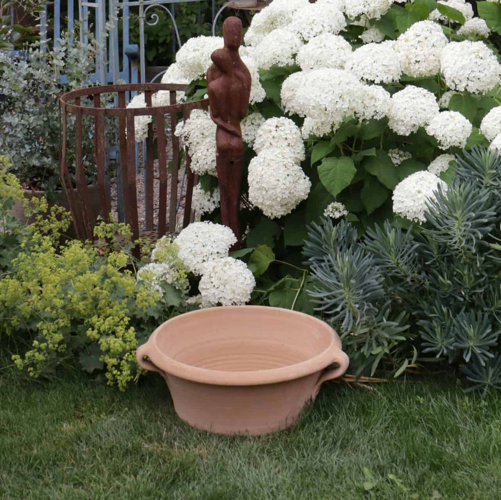 Eine Gartenszene mit der Kreta Keramik Terrakotta-Pflanzschale „Krokus“ mit einem Durchmesser von 50 cm, platziert auf grünem Gras und umgeben von üppigen Pflanzen und weißen Hortensienblüten. Eine hohe, abstrakte Holzstatue steht zwischen dem Laub, während Metallgartendekor und blühende Krokusse dem Hintergrund Charme verleihen.