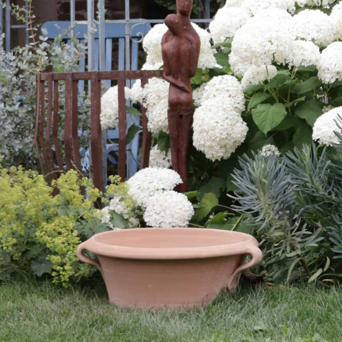 Eine Gartenszene mit der Kreta Keramik Terrakotta-Pflanzschale „Crocus“ auf grünem Gras, weißen Hortensienblüten im Hintergrund und einem verrosteten Metallstuhl. Eine hohe, rostfarbene Skulptur einer abstrakten menschlichen Figur steht inmitten der Blumen, ergänzt durch leuchtende Krokusblüten, die hindurchscheinen.
