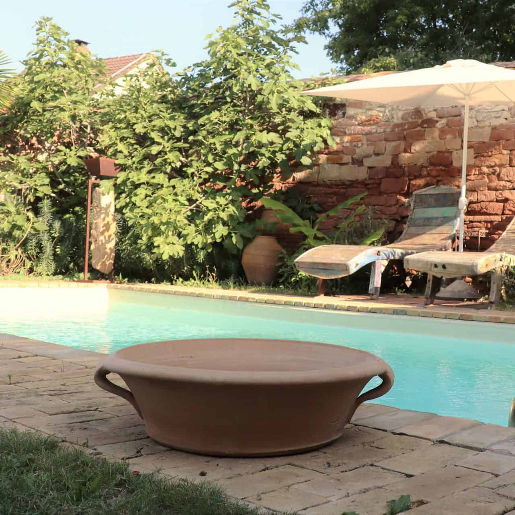 Eine ruhige Poolszene im Hinterhof mit zwei Liegestühlen unter einem großen weißen Sonnenschirm auf der rechten Seite. Eine große, flache Terrakotta-Pflanzschale „Crocus“ mit 40 cm Durchmesser steht auf dem Steinpflaster am Poolrand. Üppiges Grün, darunter leuchtende Krokusblüten und ein kleiner Baum, umgibt den Poolbereich und bietet Privatsphäre.