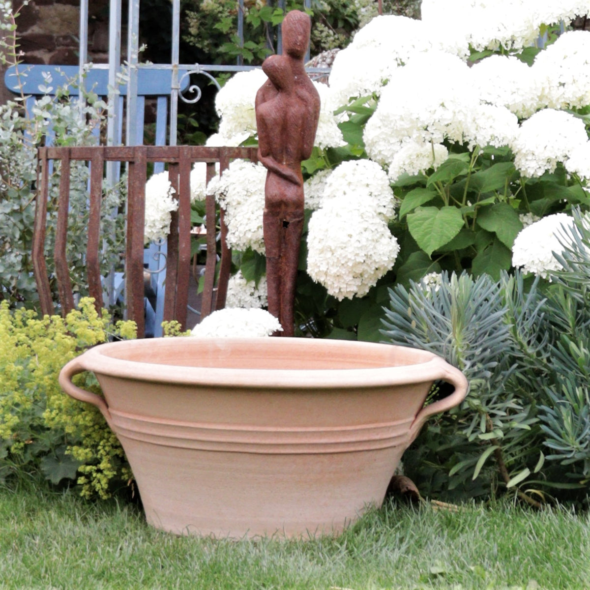 Eine Gartenszene mit einer großen Kreta Keramik Terrakotta Pflanz- und Wasserschale „Krokus“, B=70 cm, mit zwei Henkeln auf dem Gras. Hinter dem Pflanzgefäß stehen weiße Hortensien und verschiedene Grünpflanzen in voller Blüte, und eine verrostete Metallskulptur einer abstrakten menschlichen Figur ist sichtbar. Im Hintergrund ist ein blaues Metalltor zu sehen.
