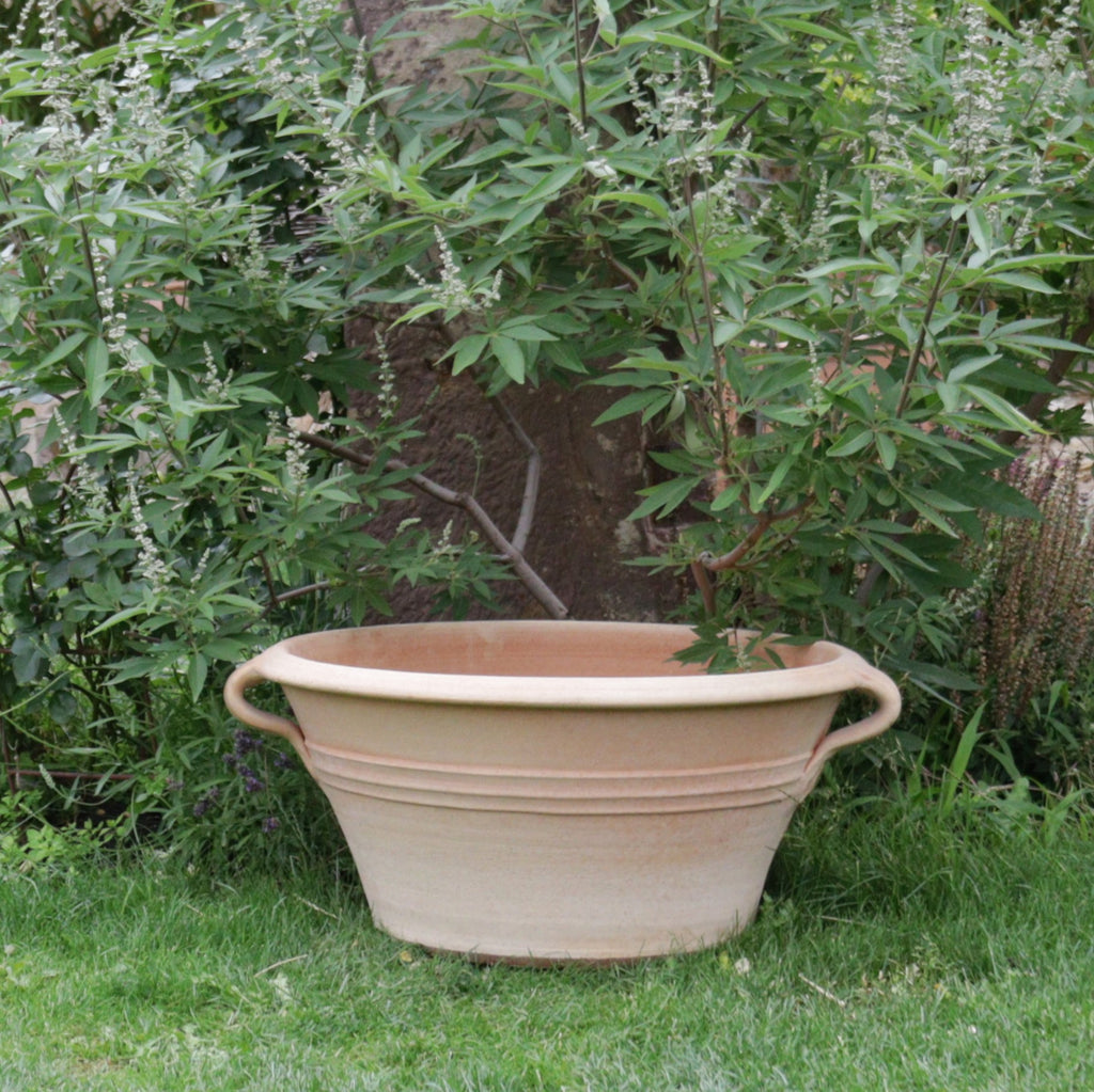 Eine große beige Kreta Keramik Terrakotta Pflanz- und Wasserschale „Crocus“ mit einem Durchmesser von 70 cm und zwei seitlichen Griffen steht leer auf dem Rasen. Dieses elegante Stück ist von üppigen grünen Pflanzen und Blättern in einer ruhigen Gartenumgebung umgeben.