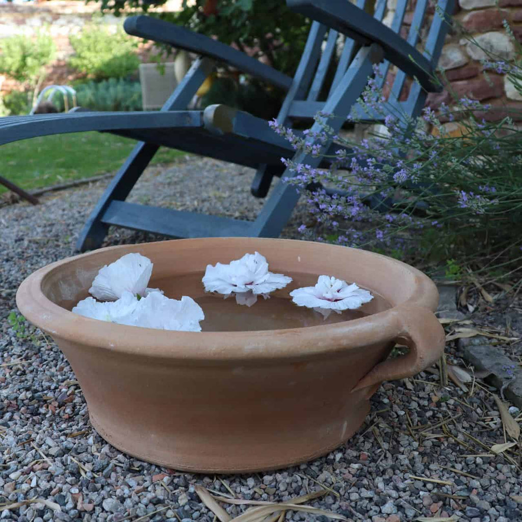 Eine Terrakotta-Pflanzschale „Krokus“ mit 40 cm Durchmesser, gefüllt mit Wasser und auf der Oberfläche schwimmen weiße Blüten, steht auf einem Kiesweg. Im Hintergrund ist ein blauer Schaukelstuhl aus Holz teilweise sichtbar, begleitet von blühenden Krokuspflanzen und üppigem Gartengrün.
