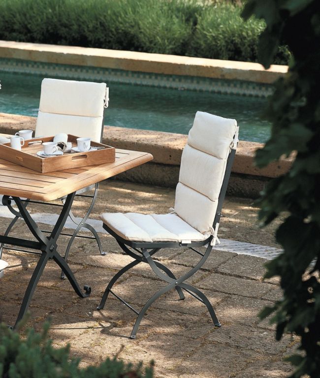 Eine ruhige Szene auf einer Außenterrasse mit einem Holztisch, auf dem ein Tablett und Teetassen stehen, umgeben von gepolsterten Klappstühlen des Typs Aurora. Im Hintergrund ist ein Swimmingpool mit klarem, blauem Wasser zu sehen, und üppiges grünes Laub umrahmt die Kulisse und sorgt für eine friedliche Atmosphäre, die an ein Polarlicht erinnert.