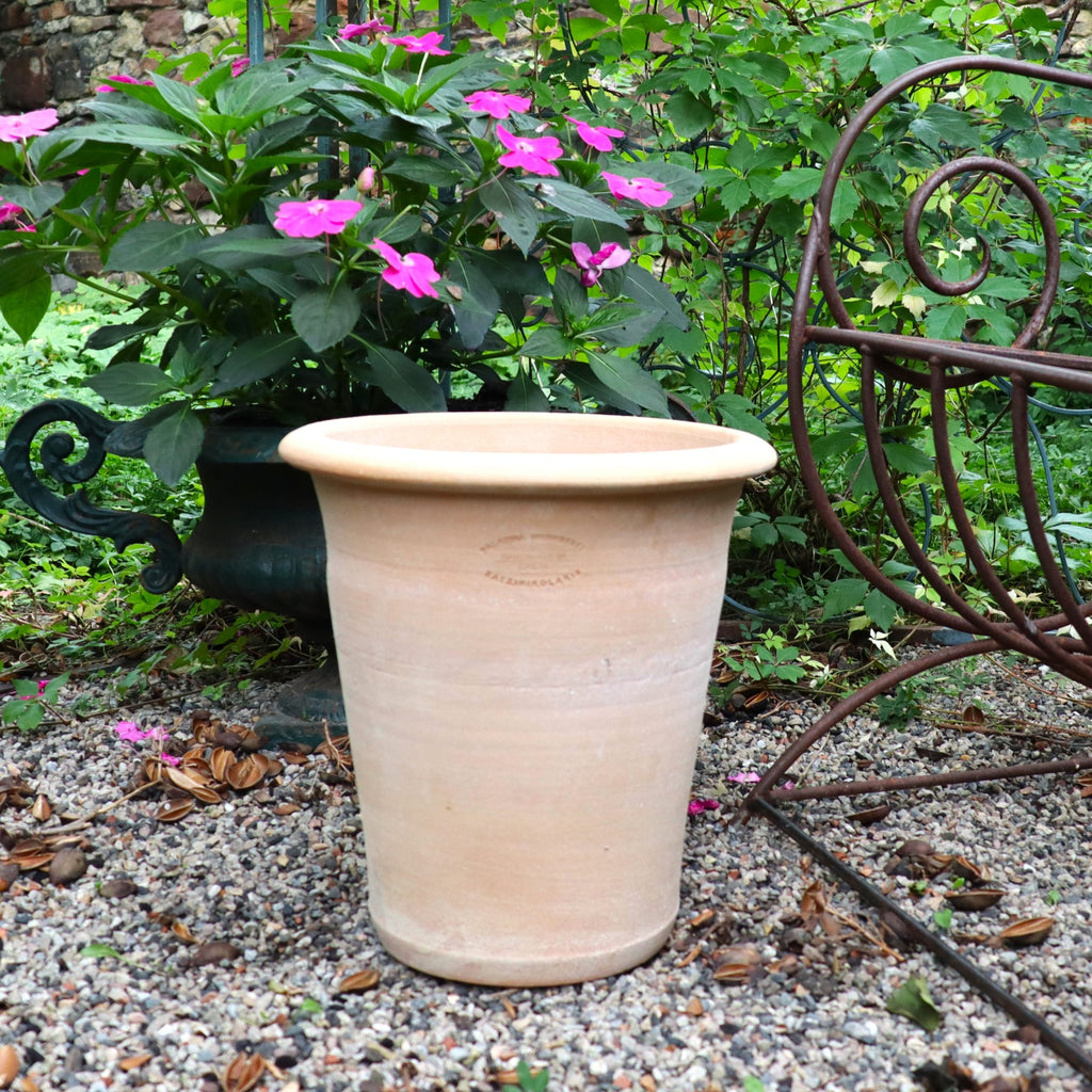 Ein großer, leerer Terrakotta-Pflanztopf "Dahlia" 35 cm - Handgemacht & Frostfest sitzt auf Gartenkies neben einer grünen Pflanze mit rosa Blüten und einem verzierten Metallstuhl, umgeben von üppigem Grün und verstreuten rosa Blütenblättern.