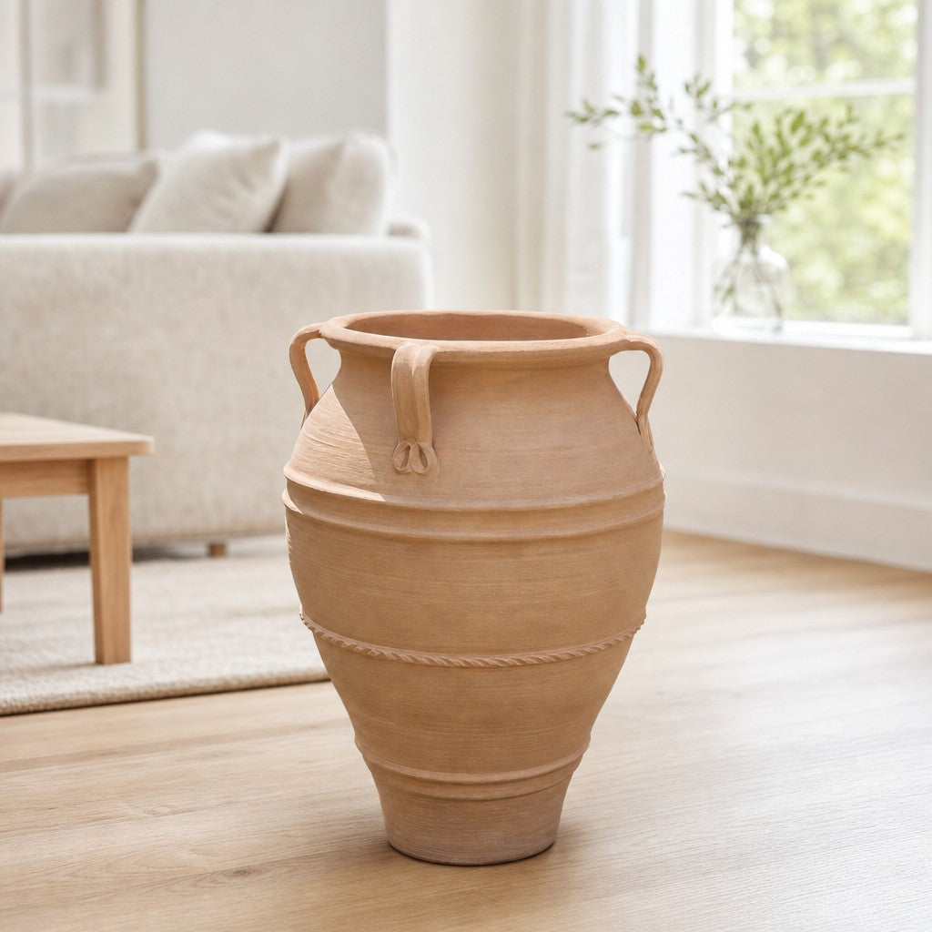 Eine XXL-Terrakotta-Amphore „Datura“ (H = 100 cm) mit eleganten Henkeln steht auf einem Holzboden in einem hellen, modernen Wohnzimmer in der Nähe eines Fensters und eines weißen Sofas, im Hintergrund ist eine kleine grüne Pflanze zu sehen.