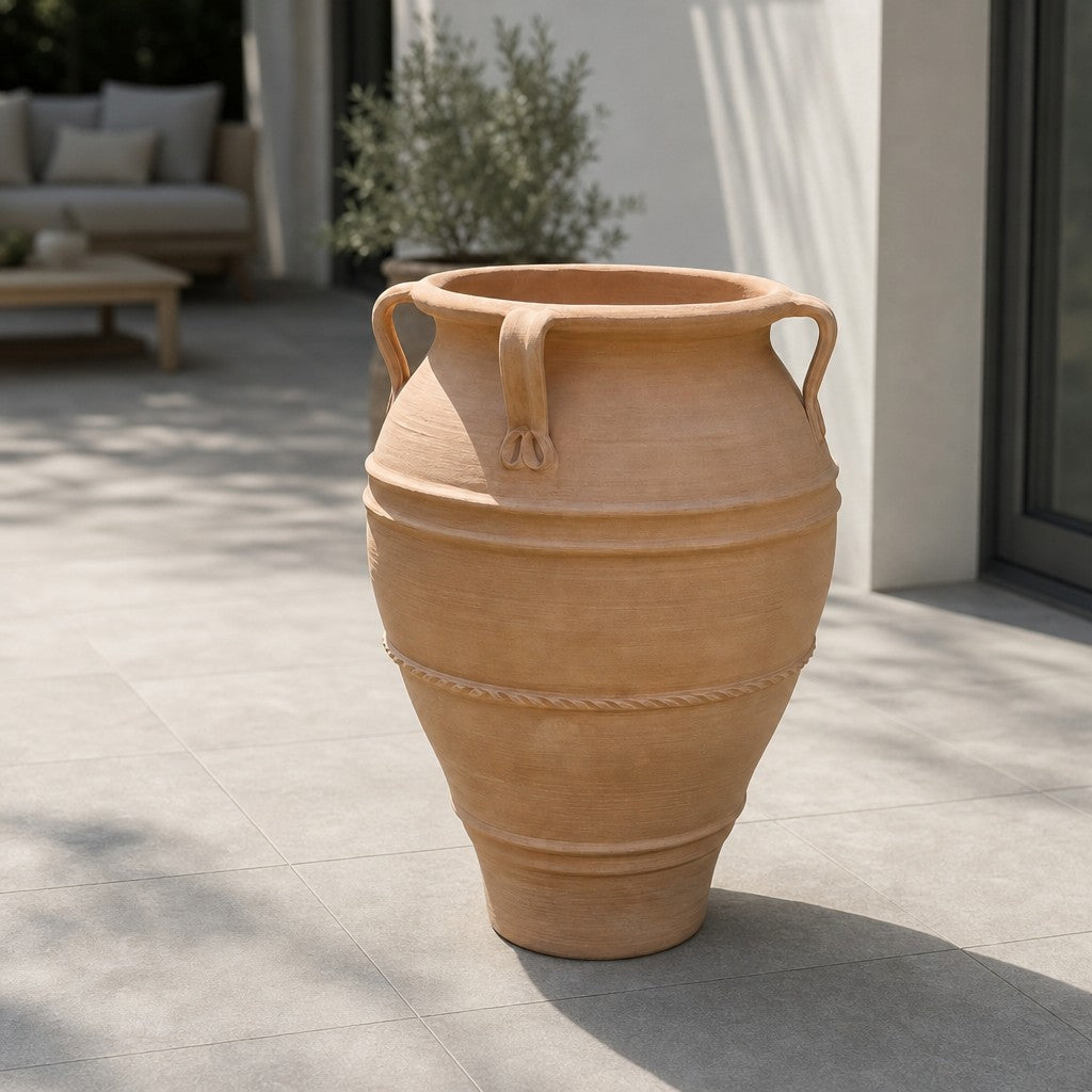 Eine XXL-Terrakotta-Amphore „Datura“ mit zwei Henkeln und einer Höhe von 100 cm steht auf einer gefliesten Terrasse neben einem modernen Gebäude; im Hintergrund sind Gartenmöbel und eine Topfpflanze zu sehen.