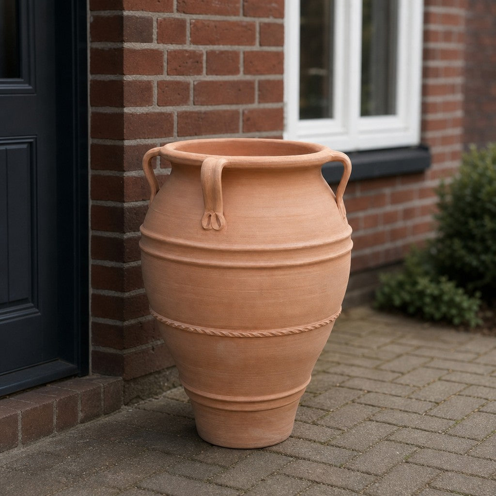 Eine XXL-Amphore aus Terrakotta mit dem Namen „Datura“ (Höhe: 100 cm) und dekorativen Henkeln steht auf einer gepflasterten Fläche neben einem Backsteingebäude mit einem weiß gerahmten Fenster.
