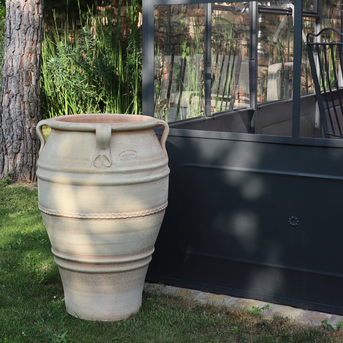 Die frostfeste Terrakotta-Amphore "Datura", H=70 cm, mit zwei Griffen steht auf grünem Gras neben einer Struktur aus schwarzem Metall und Glas, umgeben von Bäumen, Pflanzen und blühenden Datura im Hintergrund.