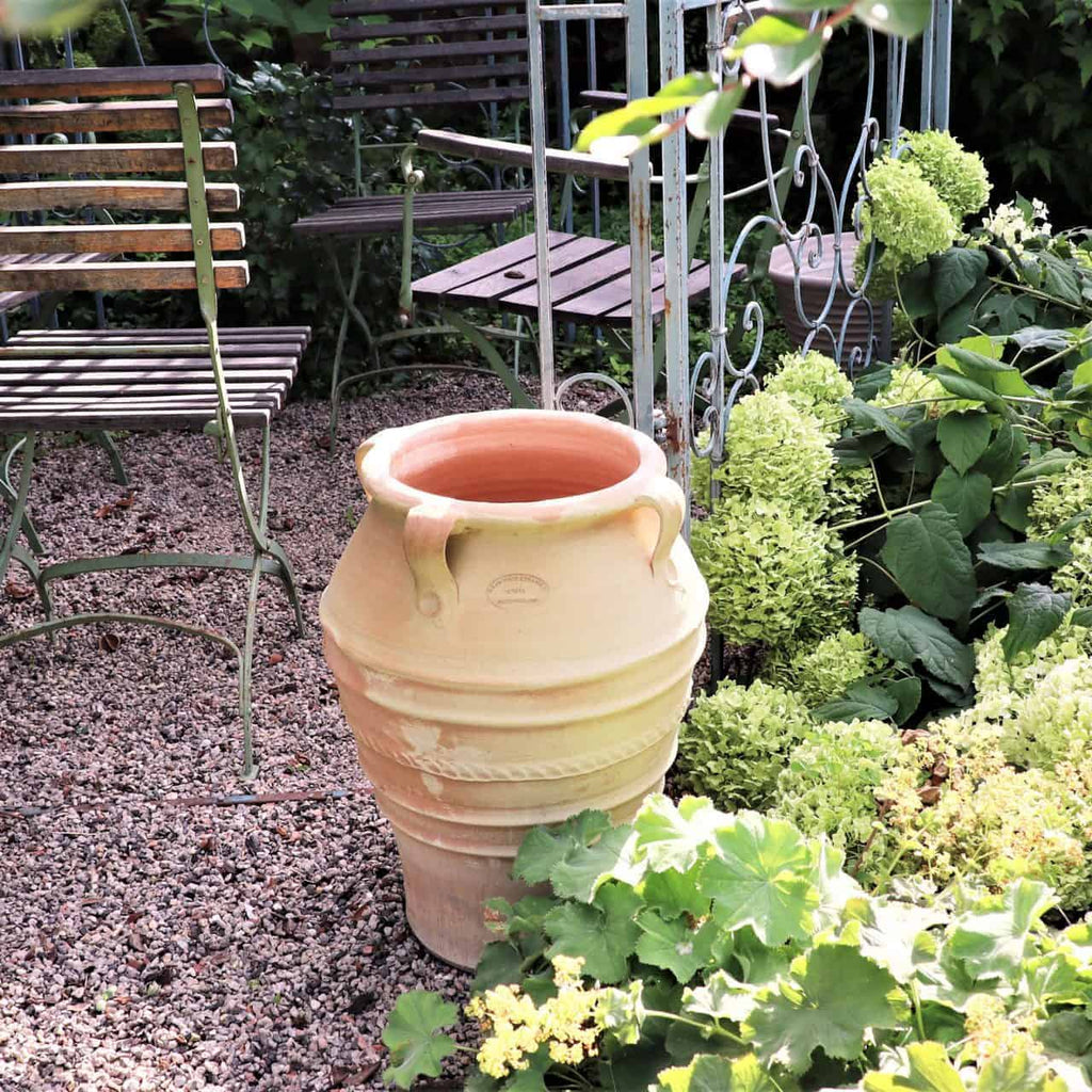 Eine Tonurne steht zwischen blühenden grünen Pflanzen in einem Garten. Um die Urne herum stehen Gartenstühle aus Metall und ein Tisch auf einer Kiesfläche. Die Szene ist üppig und ruhig, wobei das Grün einen Kontrast zum erdigen Ton der Urne bildet.