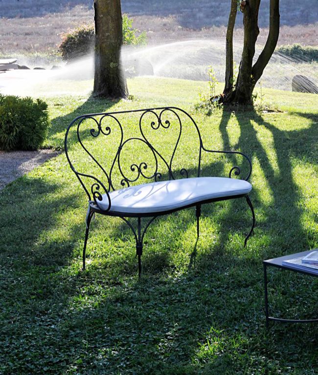 Die Sitzbank Aurora, eine schmiedeeiserne Bank mit gepolsterter Sitzfläche, steht auf einer Rasenfläche. Wassersprinkler erzeugen im Hintergrund einen sanften Nebel, während das Sonnenlicht Schatten von Bäumen und Grünflächen wirft, die an die Morgendämmerung erinnern. In der Nähe steht ein kleiner Tisch mit einem Buch oder einer Zeitschrift.