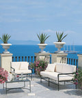 Eine Terrasse mit malerischem Meerblick, ausgestattet mit dem eleganten Sofa Toscana aus Eisen in Graphitgrau, mit plüschigen weißen Kissen und einem eleganten Couchtisch mit Glasplatte. Topfpflanzen und blühende Blumen schmücken den Raum, während in der Ferne ein Segelboot und der bergige Horizont der Toskana unter einem klaren blauen Himmel sichtbar sind.