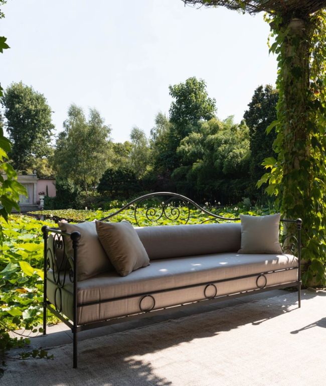 Eine elegante Gartenbank, das Sofa Aurora aus Eisen in Graphitgrau, mit aufwendigen Details und glatten Kissen, steht unter einer Pergola, die mit üppigen grünen Weinreben bewachsen ist. Von der Bank aus blickt man auf einen ruhigen Garten mit großen grünen Bäumen und einem Teich voller Seerosen im Hintergrund.