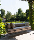 Eine elegante Gartenbank, das Sofa Aurora aus Eisen in Graphitgrau, mit aufwendigen Details und glatten Kissen, steht unter einer Pergola, die mit üppigen grünen Weinreben bewachsen ist. Von der Bank aus blickt man auf einen ruhigen Garten mit großen grünen Bäumen und einem Teich voller Seerosen im Hintergrund.