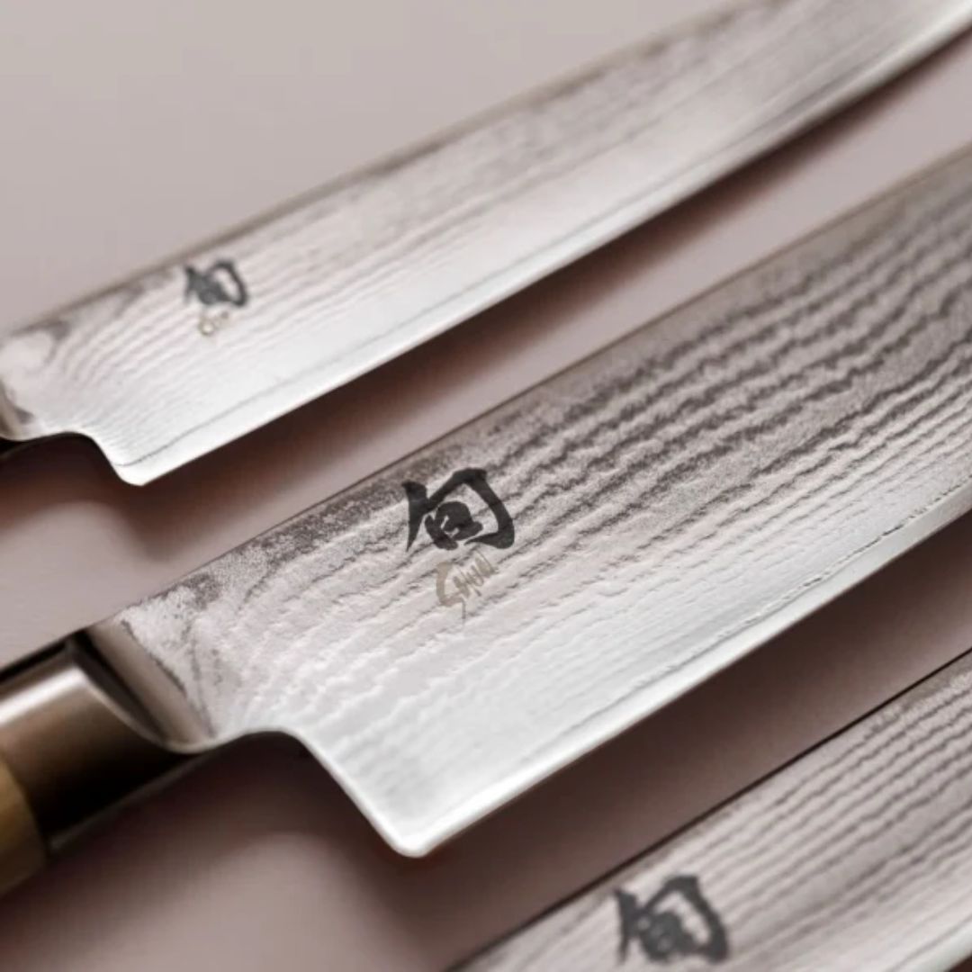 Nahaufnahme von drei KAI Shun Classic DM-0702W Santokumessern mit 18 cm langen Damaststahlklingen und Kanji-Gravuren, die diagonal auf einer hellen Fläche angeordnet sind.