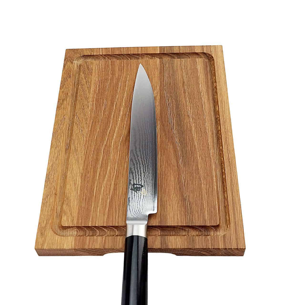 Ein scharfes Kai Shun Classic DM-0704 Schinkenmesser mit schwarzem Griff liegt auf einem 30 x 22 cm großen Schneidebrett aus Eiche. Das Brett hat eine Rille um die Kante, um den Saft aufzufangen. Die Damastklinge des Messers weist eine feine Oberfläche und ein ausgeprägtes Muster auf, was sein elegantes Design unterstreicht.