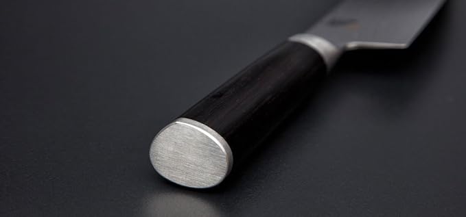 Nahaufnahme des Griffs eines Kai Shun DM-0705 Brotmessers Klinge 23,0 cm. Der Griff ist schwarz mit einer glatten, polierten Oberfläche und einer metallischen Endkappe. Die Klinge erstreckt sich unscharf im Hintergrund und kontrastiert mit der dunklen Oberfläche, auf der das Messer ruht, und wird durch ein Fassholzbrett mit den Maßen 30 x 18 cm ergänzt.