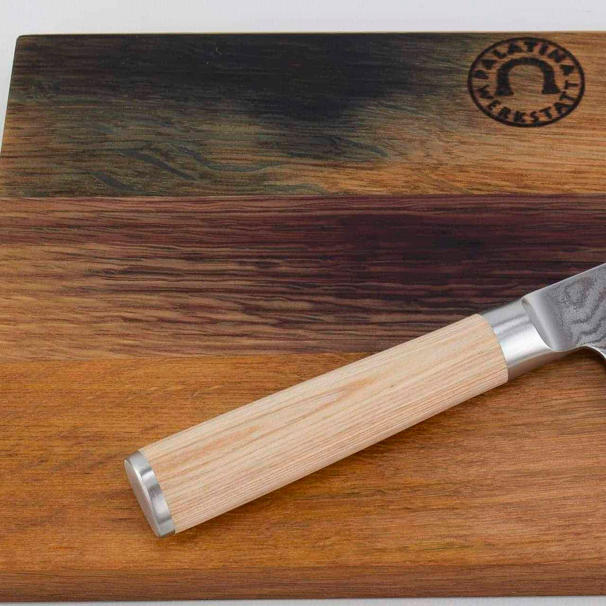 Eine Nahaufnahme eines limitierten Kiritsuke-Messers Kai Shun Classic White DM 0777W mit Holzgriff, das auf einem handgefertigten Fassholzbrett 30x18 cm ruht, das verschiedene Holzmaserungen und Farben aufweist. In der oberen rechten Ecke des Schneidebretts ist ein Logo in das Holz eingeprägt.