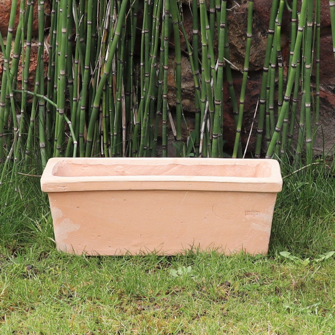 Der rechteckige Pflanzkasten Terrakotta „Erica“ L=40 cm, hellbraun in der Farbe, steht auf einer Rasenfläche mit hohen Bambuspflanzen im Hintergrund. Der Pflanzkasten ist leer und leicht verwittert, vor einer Steinmauer positioniert, die teilweise durch den Bambus verdeckt ist.