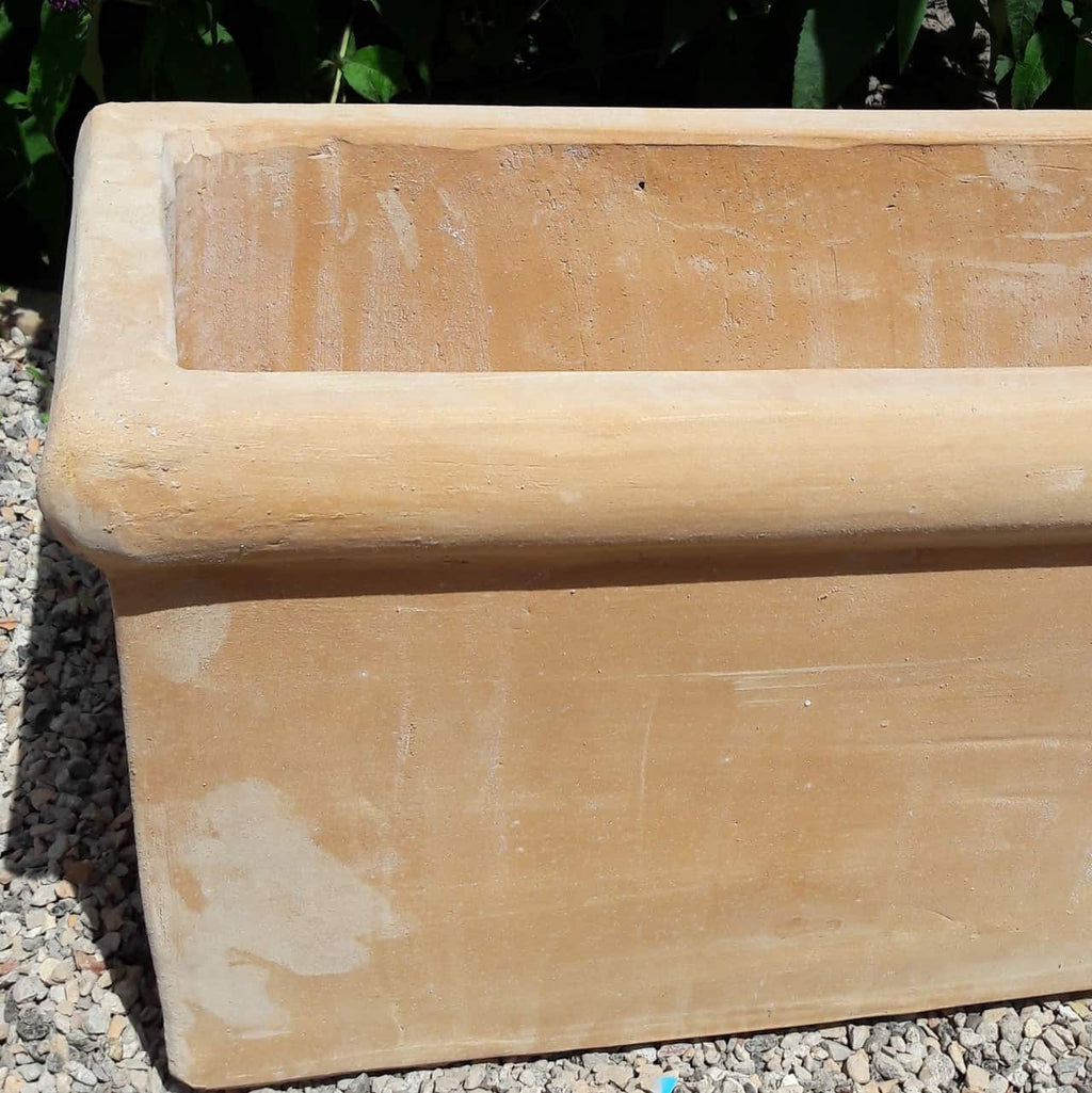Ein rechteckiger, hellbrauner Pflanzkasten Terrakotta „Erica“ L=60 cm steht auf einer Kiesfläche im Freien. Der Pflanzkasten hat einen breiten Rand und scheint verwittert zu sein, mit sichtbaren Kratzspuren und leichter Verfärbung. Im Hintergrund ist leicht grünes Laub zu sehen, das dem rechteckigen Pflanzkasten einen Kontrast verleiht.