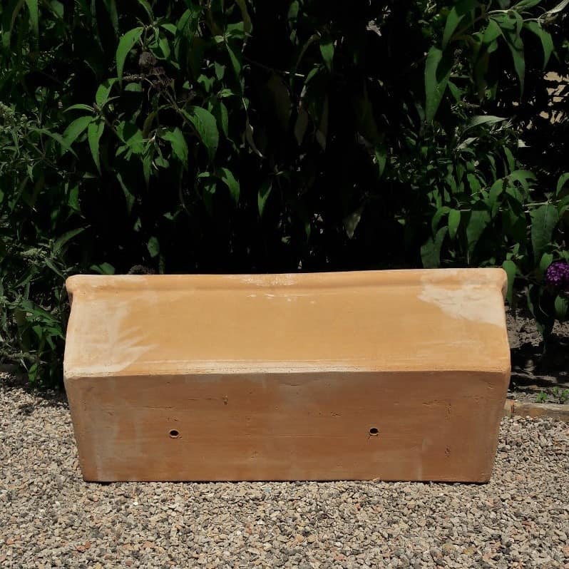 Der rechteckige Pflanzkasten Terrakotta „Erica“ L=60 cm steht auf einer Kiesfläche. Am Boden befinden sich kleine Drainagelöcher. Hinter dem Pflanzkasten bildet üppiges grünes Laub einen kontrastreichen Hintergrund. Der Pflanzkasten ist leer und dem Sonnenlicht ausgesetzt, bereit für leuchtende Erica-Blüten.