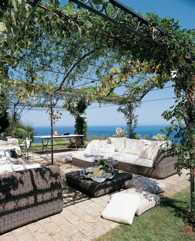 Ein gemütlicher Sitzbereich im Freien mit Korbmöbeln und weißen Kissen befindet sich unter einer freistehenden Pergola Tibisco mit Überdachung, die mit Grünpflanzen behangen ist. Der Raum bietet einen malerischen Blick auf das Meer. Ein niedriger Tisch in der Mitte bietet Platz für Getränke und Dekoration, während zusätzliche Kissen und Polster zu einer entspannten Atmosphäre beitragen.