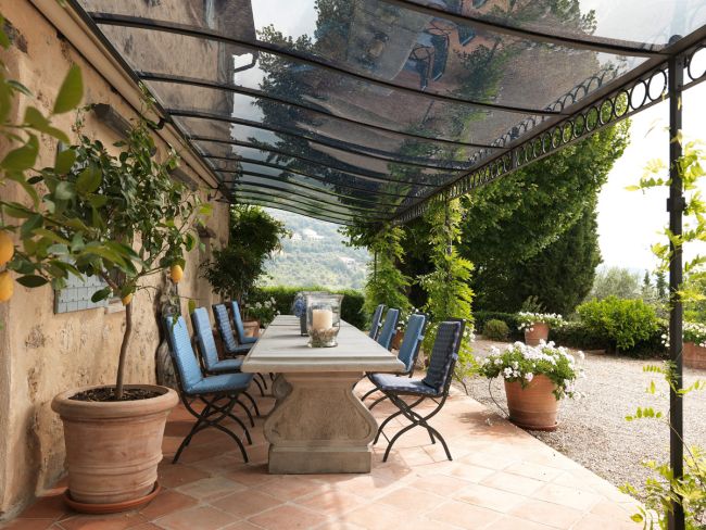Ein Essbereich im Freien verfügt über einen langen Tisch mit blau gepolsterten Stühlen auf einer mit Terrakottafliesen ausgelegten Terrasse. Der Raum wird von einer Anbaupergola Tibisco mit Überdachung aus Polykarbonat beschattet und ist von Topfpflanzen umgeben. Er bietet einen malerischen Blick auf das Grün und die Hügel im Hintergrund.