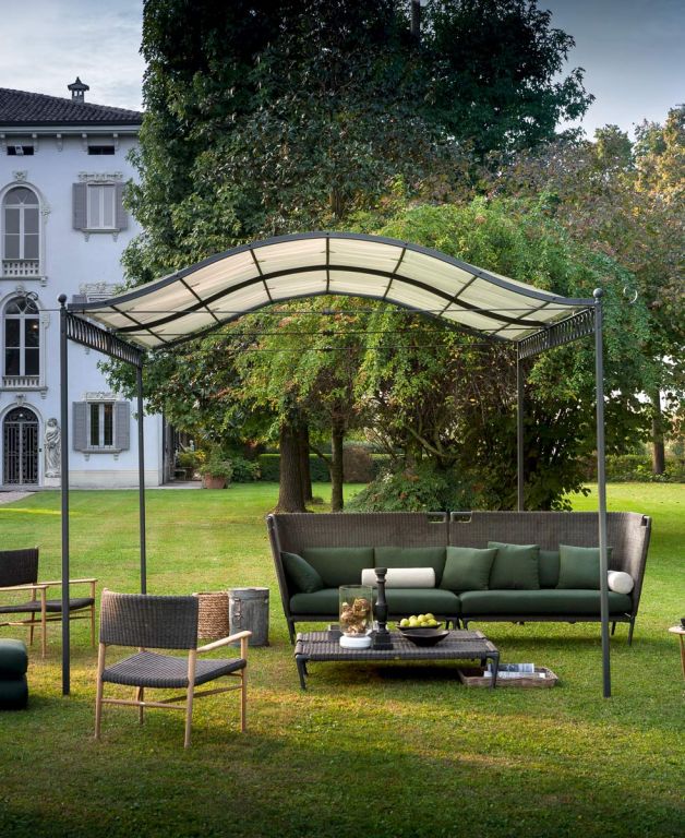 Ein ruhiger Loungebereich im Freien zeichnet sich durch die freistehende Pergola Tibisco mit Überdachung aus, die mit ihrem eleganten, geschwungenen, transparenten Design aufwartet. Darunter befinden sich Korbmöbelstücke von Tibisco, darunter ein großes Sofa mit grünen Kissen, ein kleiner Stuhl und ein Couchtisch. Die Einrichtung befindet sich auf einer Rasenfläche mit Bäumen und einem Gebäude im Hintergrund.