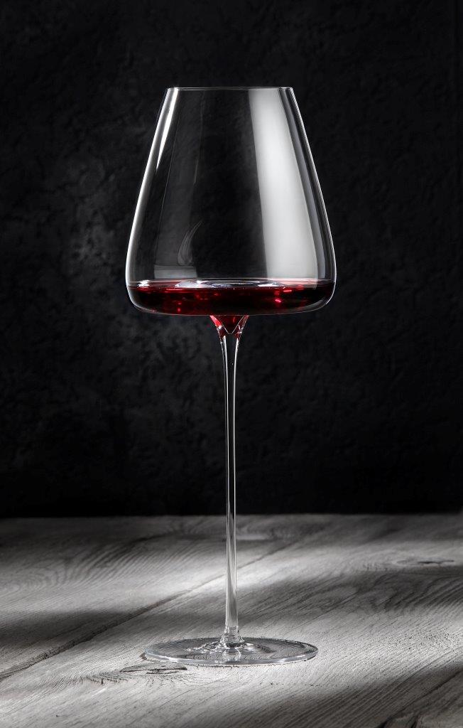 Ein Zieher Weinglas "Straight" aus der VISION-Serie, konzipiert für 100% authentischen Weingenuss, steht mit seinem dünnen Stiel, teilweise gefüllt mit Rotwein, auf einer hellen Holzfläche vor einem dunklen, strukturierten Hintergrund.