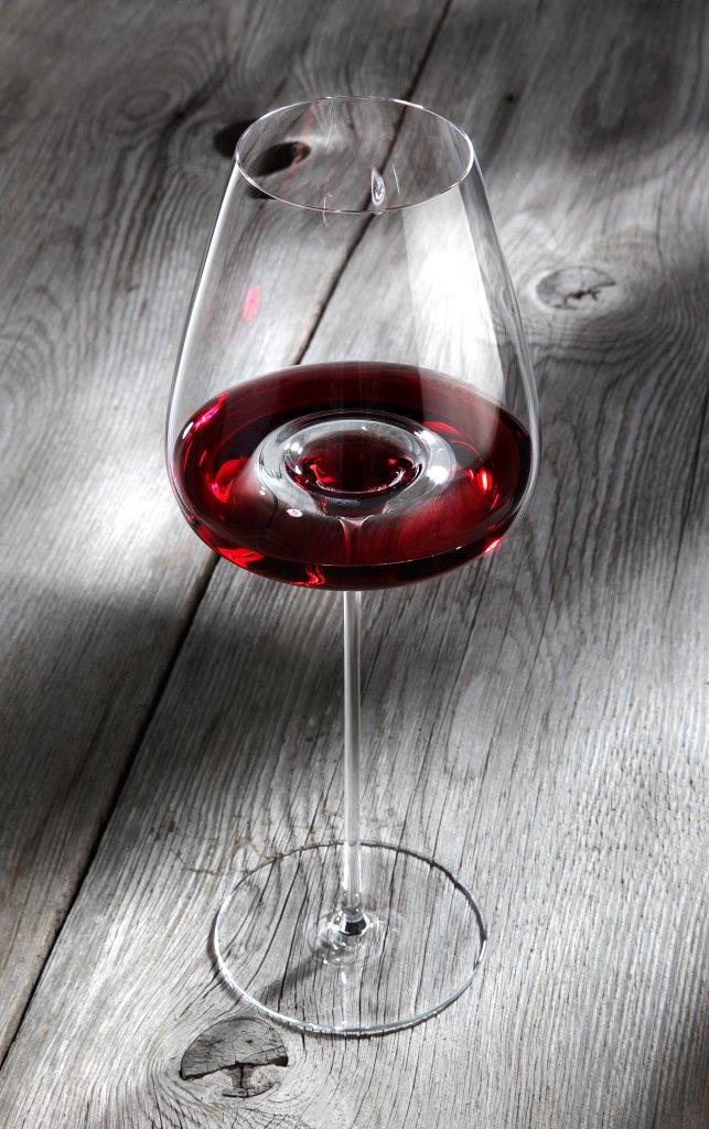 Ein Glas Rotwein ruht auf einer rustikalen Holzfläche, seine satte Farbe und die elegante Form des Zieher Weinglas "Straight" - VISION-Serie | 100% authentischer Weingenuss werden durch Licht und Schatten hervorgehoben.