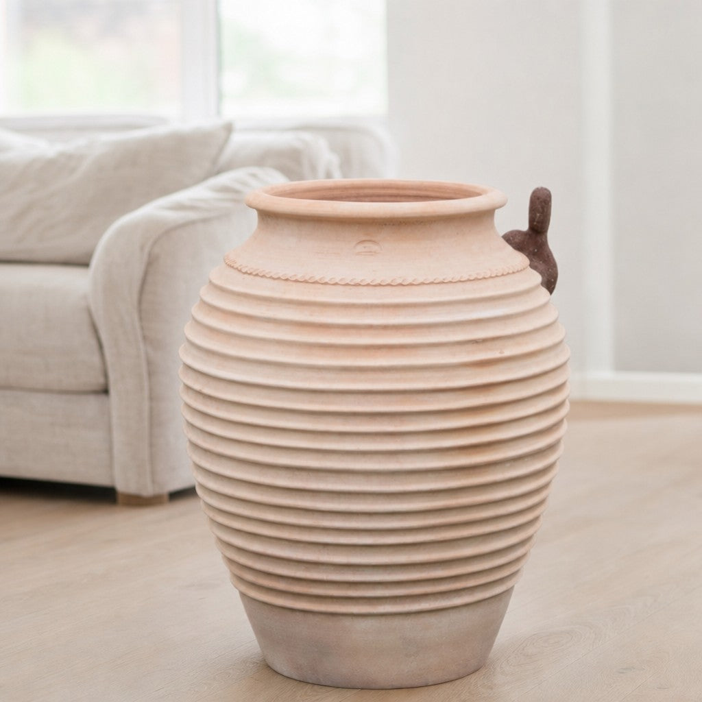 Eine große, gerippte Keramikvase, inspiriert von der winterharten Dekorationsamphore „Ficus“ (H = 100 cm), steht auf einem hellen Holzboden in einem modernen, hellen Wohnzimmer, im Hintergrund ein beiges Sofa und daneben ein hoher Ficus (80 cm).