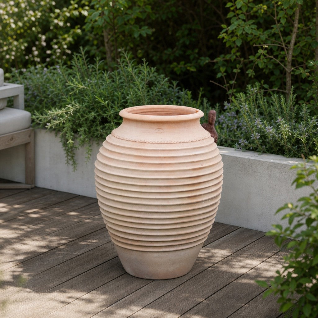 Auf einer Holzterrasse steht eine große, gerippte Terrakotta-Urne, umgeben von üppig grünen Sträuchern und Kräutern; in der Nähe sind ein weißer Beton-Pflanzkübel und ein Teil einer gepolsterten Bank zu sehen. Abgerundet wird diese elegante Kulisse durch die winterharte Dekorationsamphore „Ficus“ (H = 100 cm), die der Außenanlage sowohl Charme als auch Widerstandsfähigkeit verleiht.