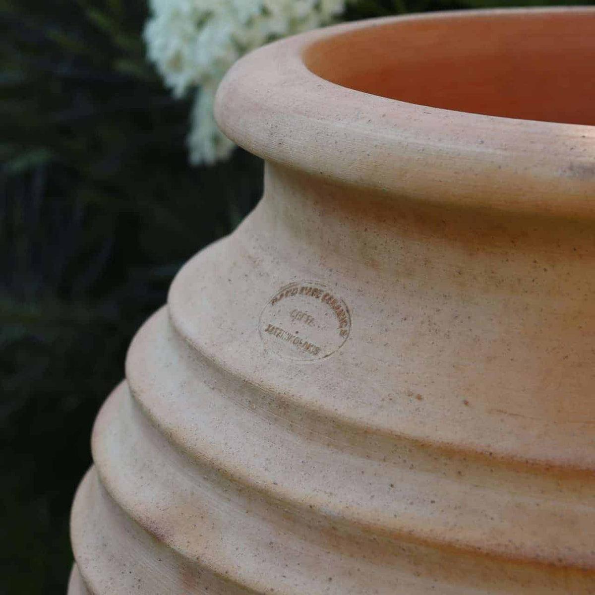 Nahaufnahme eines großen, beigen, gerippten Tontopfs. Die Terrakotta-Amphore Garten „Ficus“ H=60cm hat am Rand einen Stempel mit der Aufschrift „GERINO FINE RAMA...“. Der Hintergrund ist leicht verschwommen und zeigt einige grüne Blätter und weiße Blüten, was auf eine ruhige Gartenumgebung hindeutet.